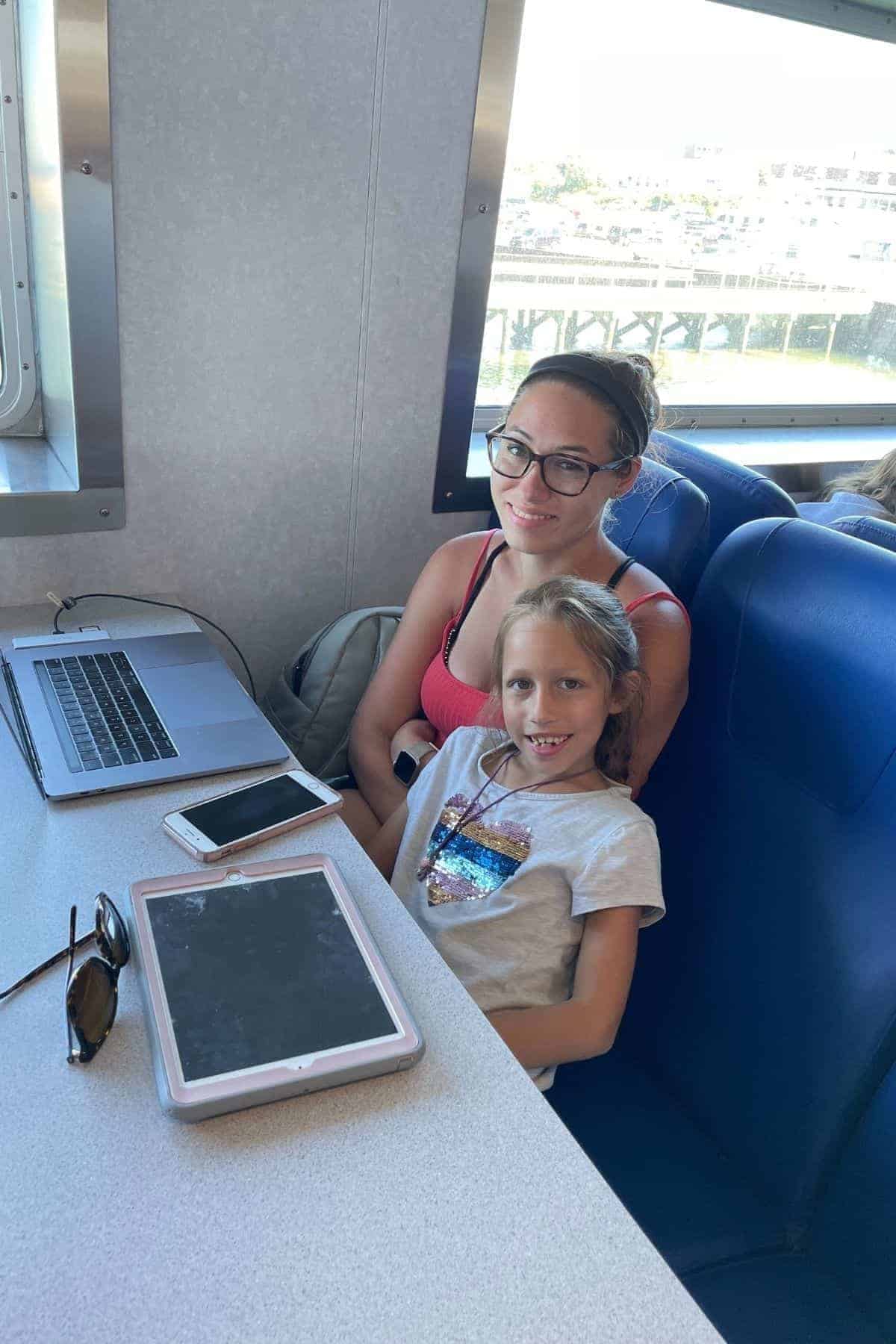 A woman and a girl sit together on a blue seat by a window, planning their Block Island day trip with a laptop, phone, and tablet on the table in front of them.