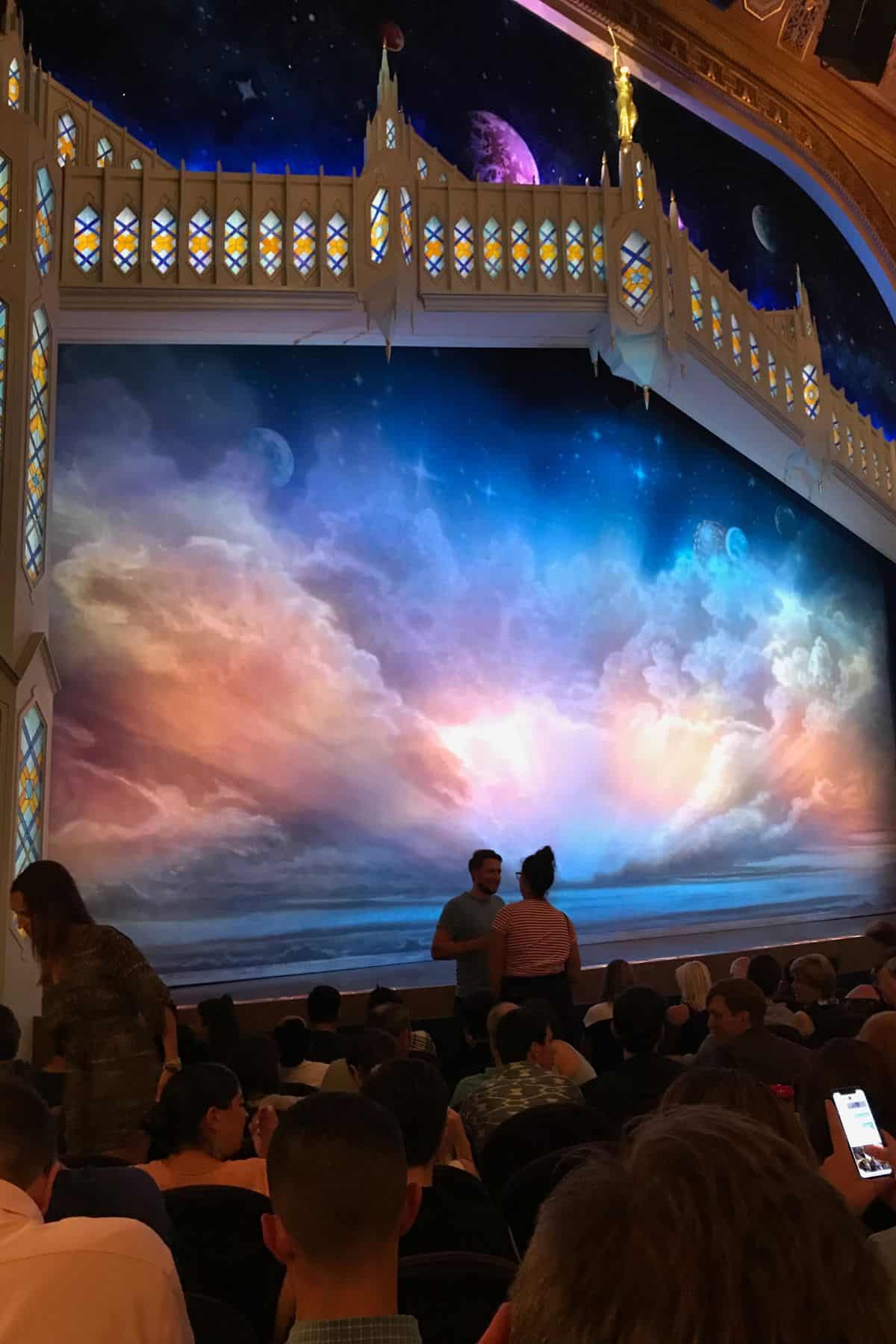 Audience seated in a theater facing a stage with a painted backdrop of clouds, sky, and planets—an enchanting scene reminiscent of a highlight from a One Day in NYC Itinerary—framed by an ornate decorative structure.