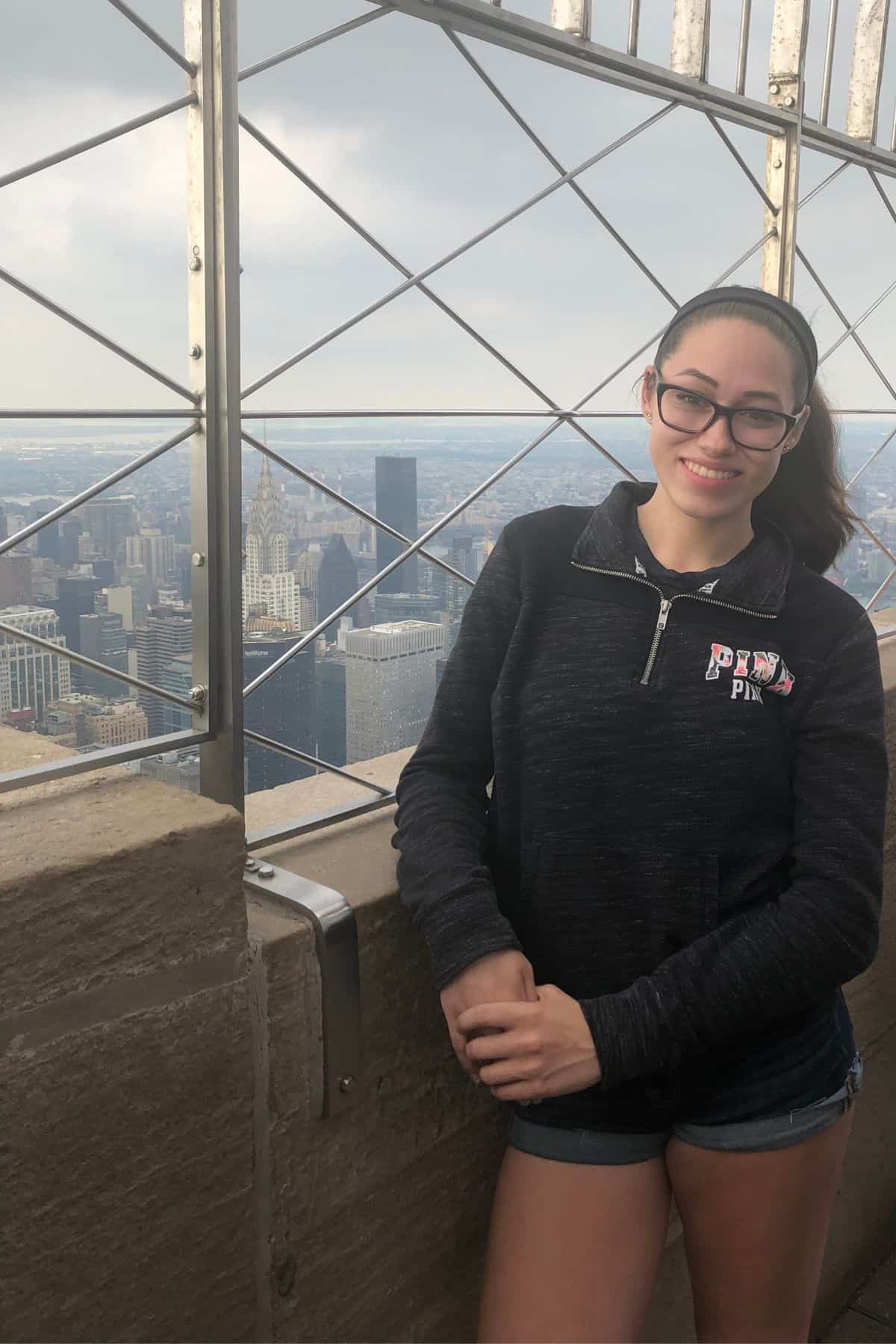 A woman wearing glasses and a black sweatshirt stands on an outdoor observation deck, taking in the city skyline under a cloudy sky—an inspiring view for anyone planning their One Day in NYC Itinerary.