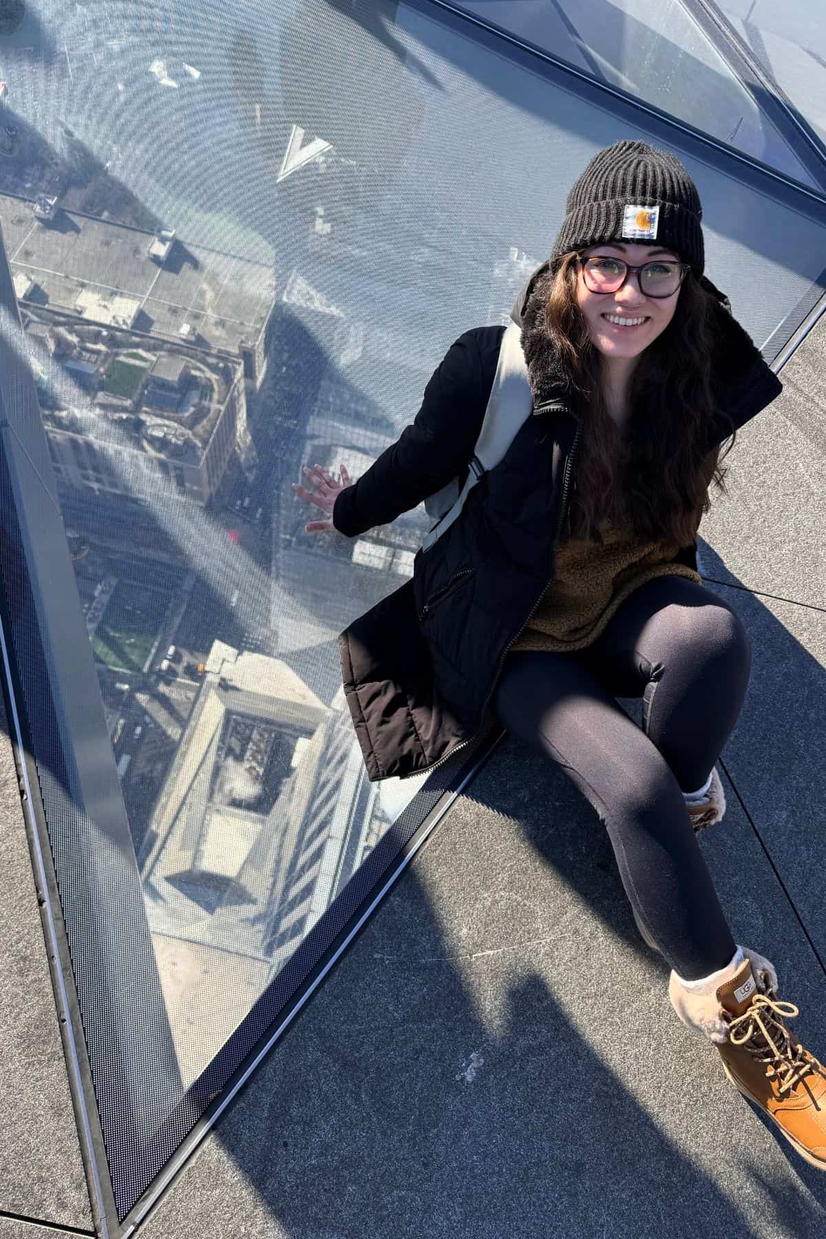 A woman in winter clothing sits on a glass floor with a city view below, smiling at the camera—capturing the perfect moment for your One Day in NYC Itinerary.