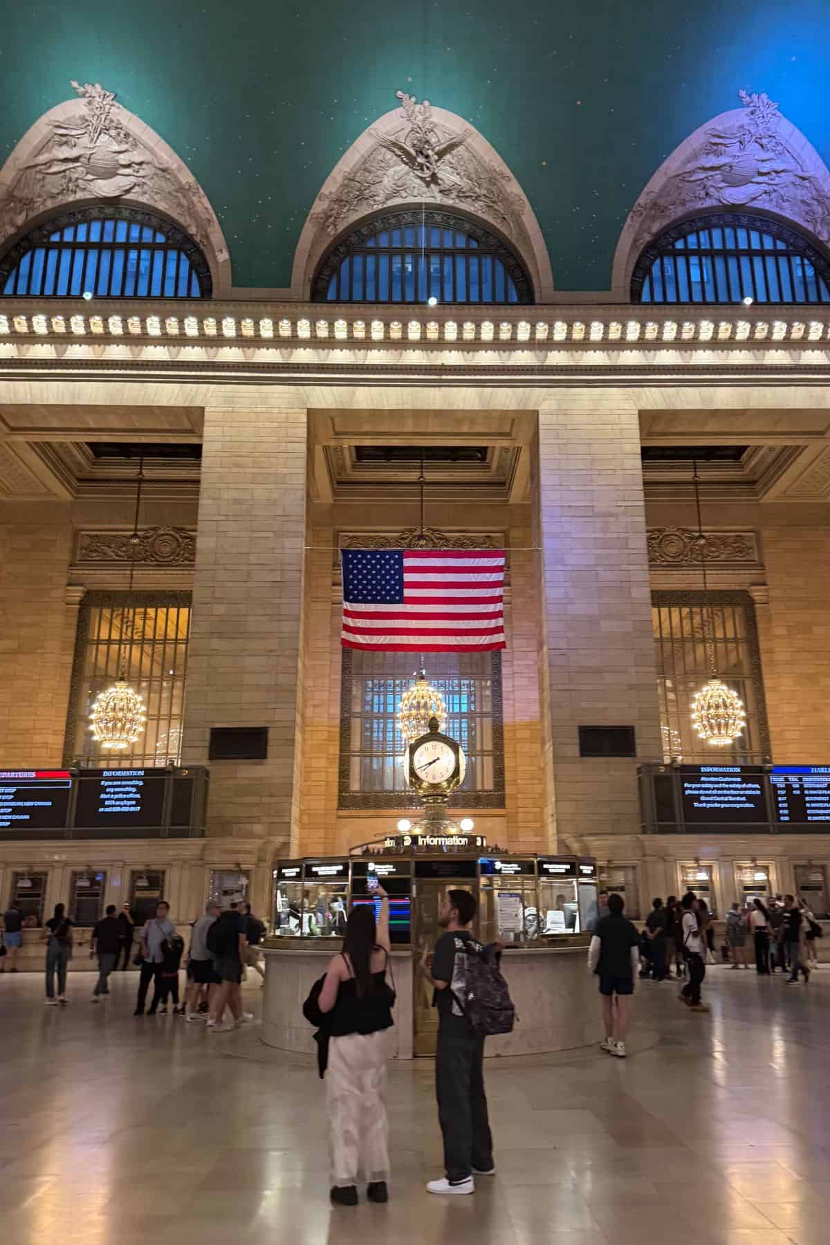 People stand in the main concourse of Grand Central Terminal, a must-see stop on any One Day in NYC Itinerary, facing an information booth with a large clock and an American flag hanging above it.