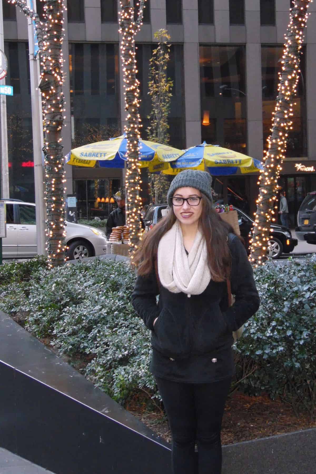 A woman wearing a gray beanie, glasses, and a scarf stands in front of decorated trees and street food umbrellas—capturing the lively spirit of a One Day in NYC Itinerary.