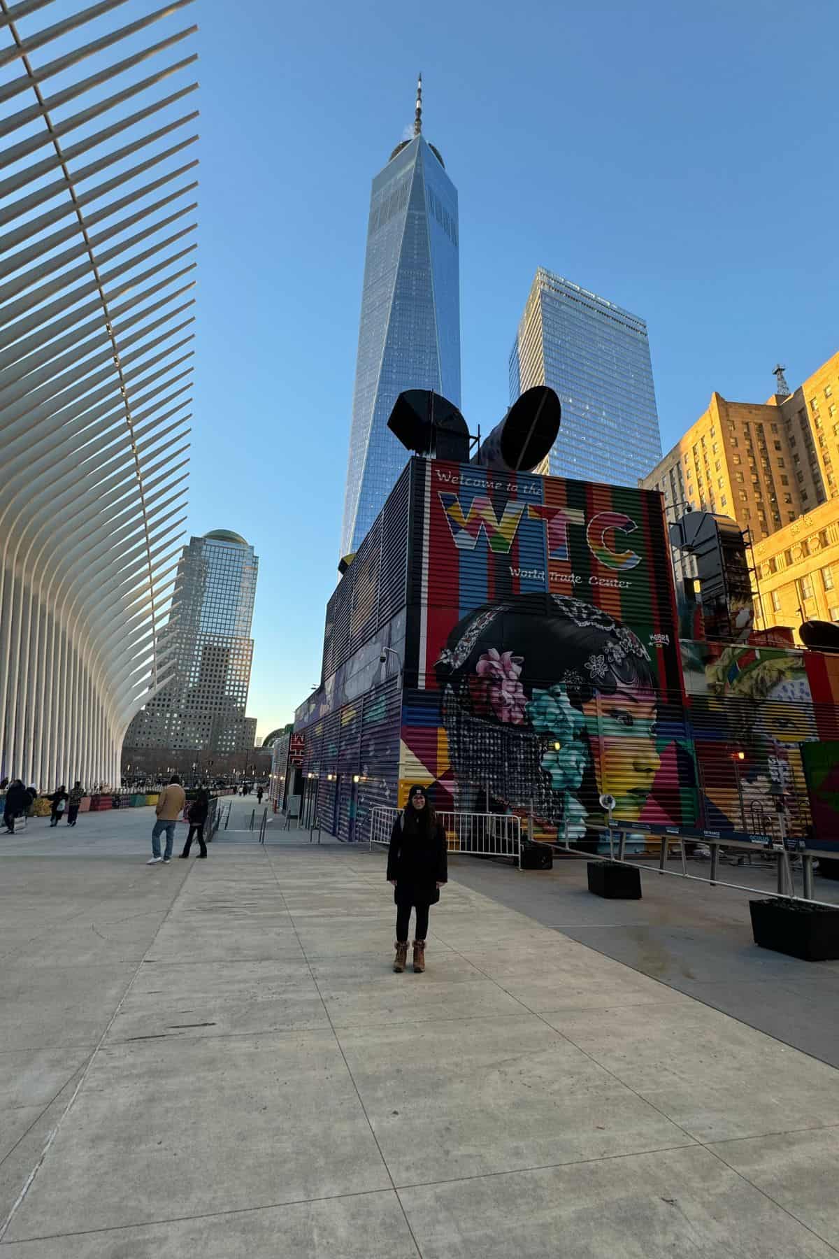 A person stands in an urban plaza with tall buildings, including One World Trade Center, and colorful murals in the background under a clear blue sky—perfect for a memorable photo stop on your One Day in NYC Itinerary.
