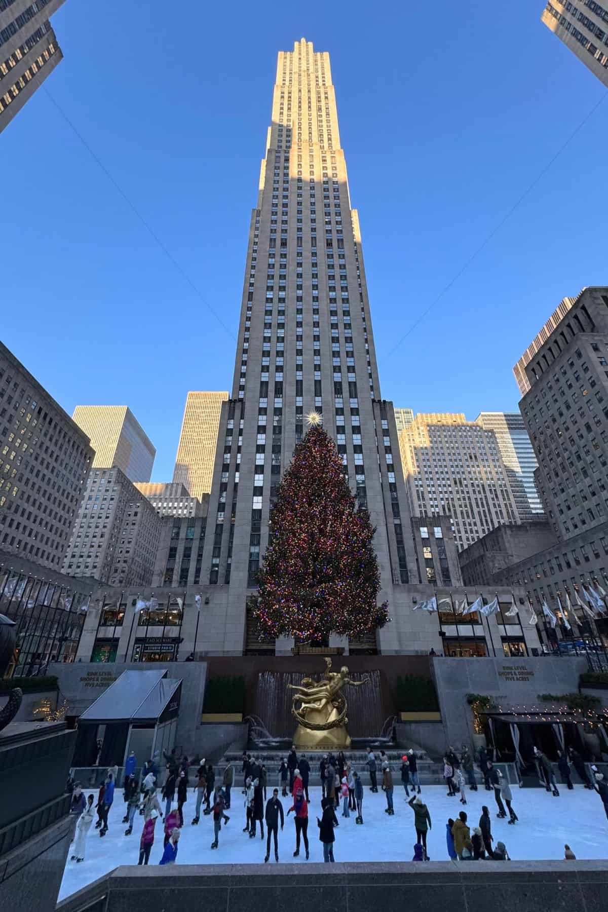 People ice skating at Rockefeller Center, a highlight for any One Day in NYC Itinerary, with a large decorated Christmas tree in front of a tall building under a clear blue sky.