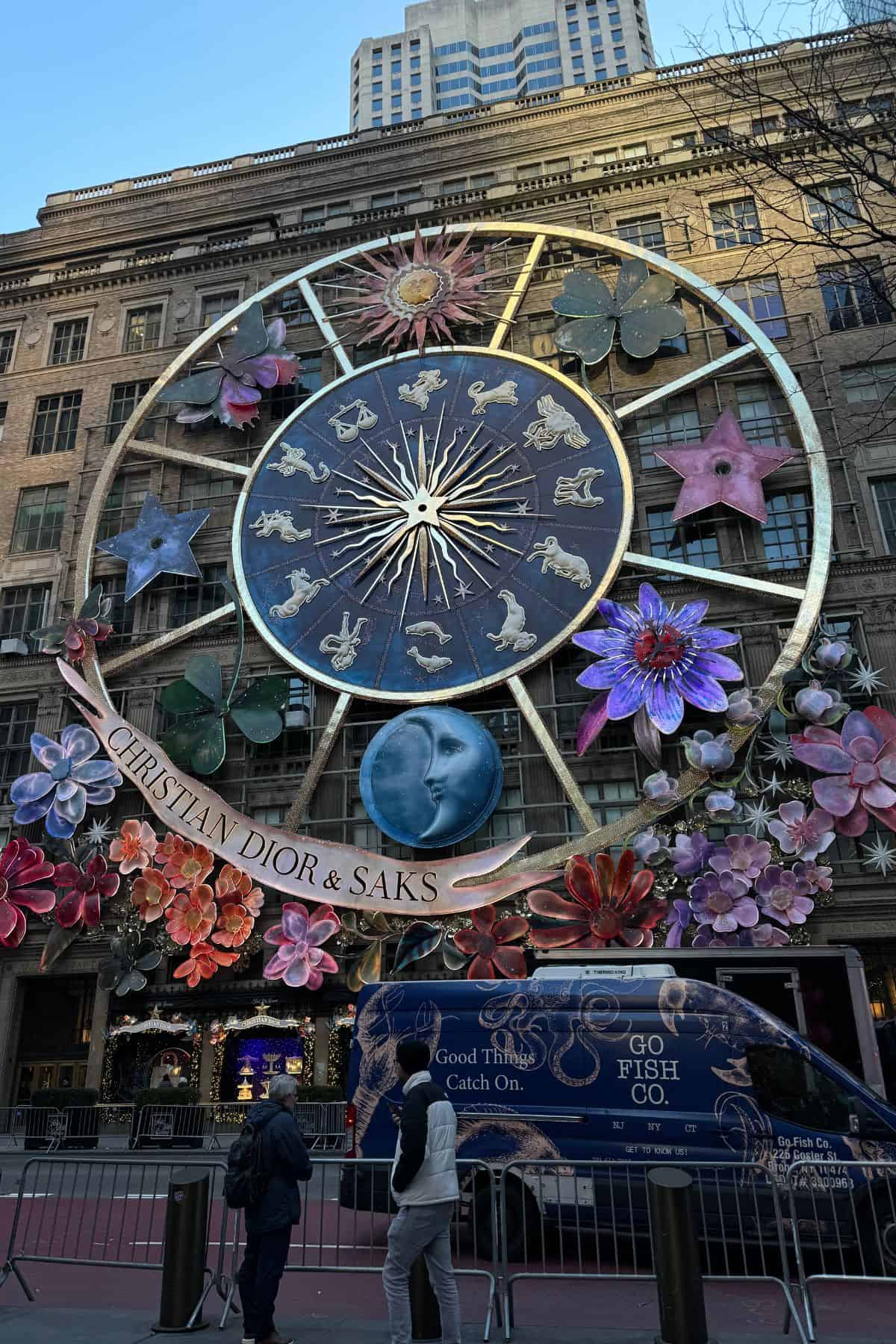 Large zodiac-themed holiday display with floral decorations and a "Christian Dior & Saks" sign on a department store facade; perfect for capturing memories on your One Day in NYC Itinerary. People and a Go Fish NYC van are in front.