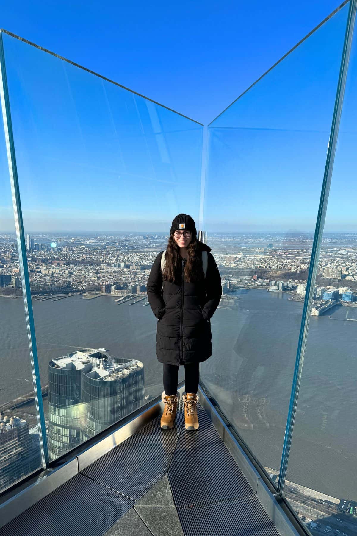 A person in a black coat, hat, and boots stands on a glass observation deck high above the city with a river in the background on a clear day—capturing an iconic moment perfect for any One Day in NYC Itinerary.