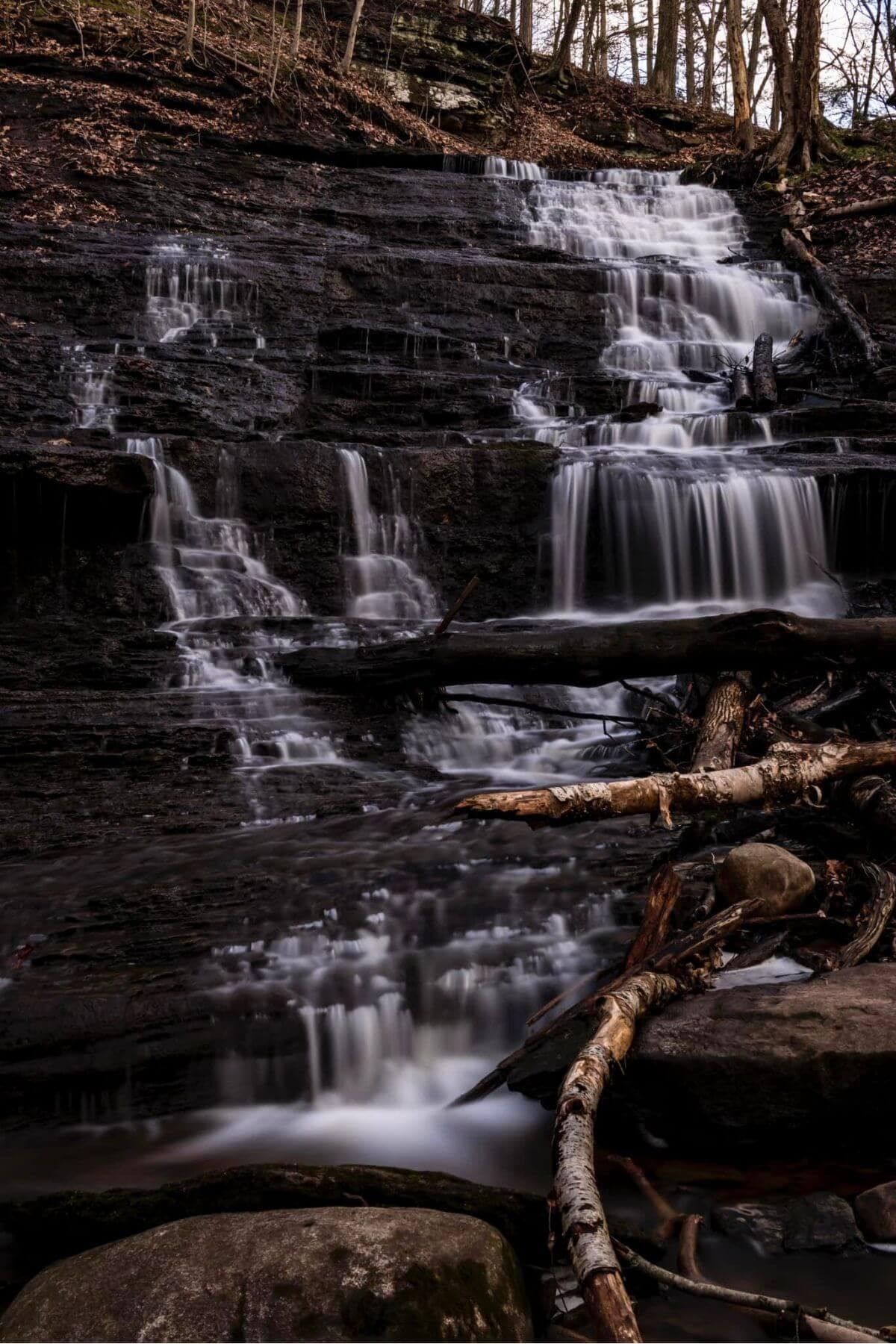 A cascading waterfall flows over rock ledges in a Connecticut forest, with fallen logs scattered across the water at its base.
