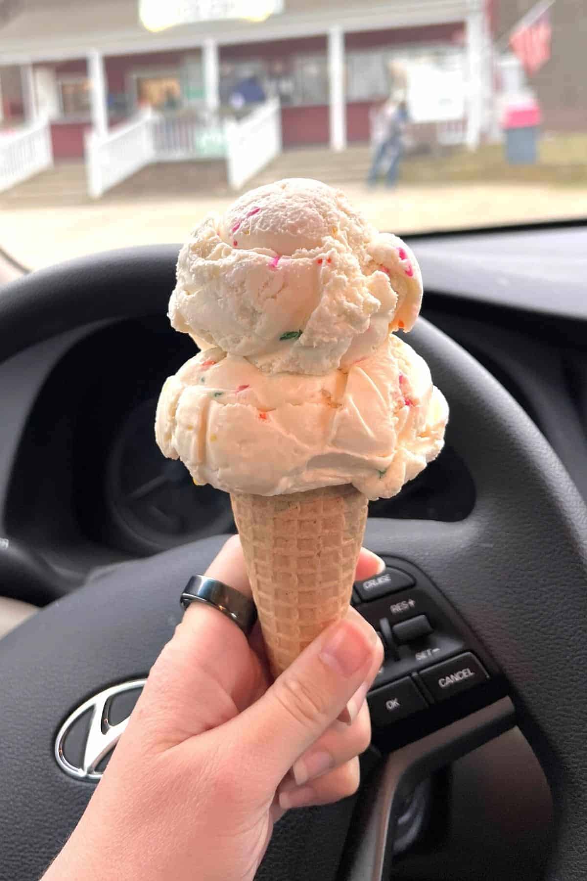 A hand holds a double scoop Salem Valley Ice Cream cone inside a car, with the steering wheel and part of the dashboard visible in the background.