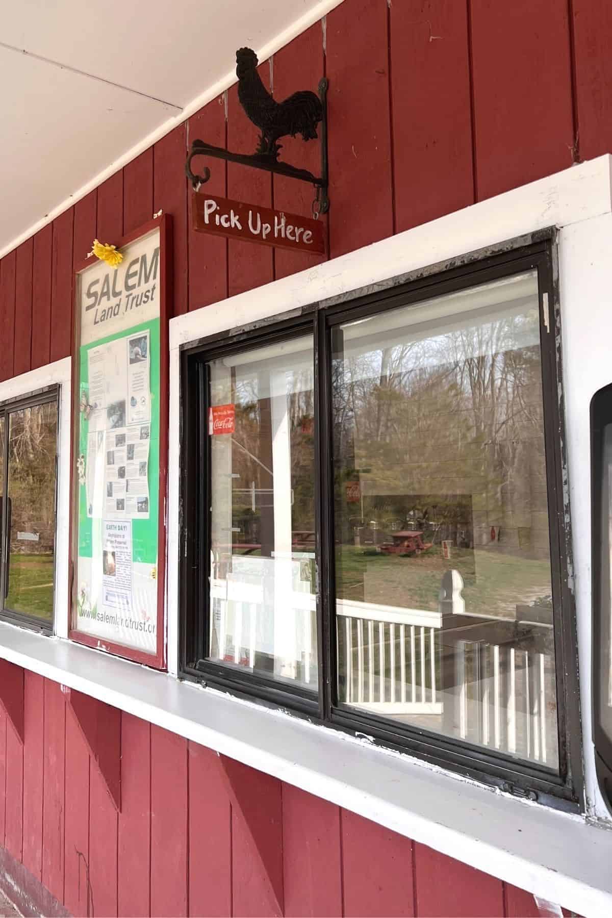 Red building with a pickup window and a sign reading "Pick Up Here" featuring a rooster silhouette; a bulletin board labeled "Salem Land Trust" is mounted on the wall—perfect backdrop for enjoying Salem Valley Ice Cream.