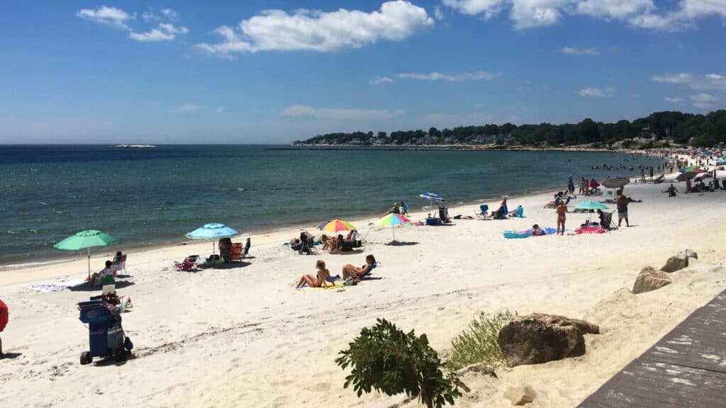 A sandy beach at Rocky Neck State Park, with scattered people sunbathing and umbrellas under a clear blue sky.