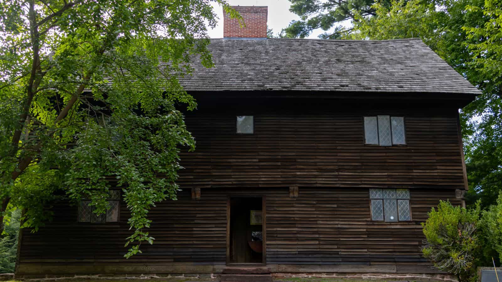 Two-story wooden house with dark horizontal siding, a shingled roof, a brick chimney, and leaded glass windows, a quintessential sight for Connecticut things to do in fall.