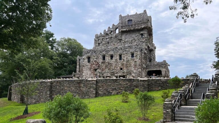 A stone castle with multiple levels and jagged edges, surrounded by greenery and a stone wall, with a staircase leading up to it under a cloudy sky.