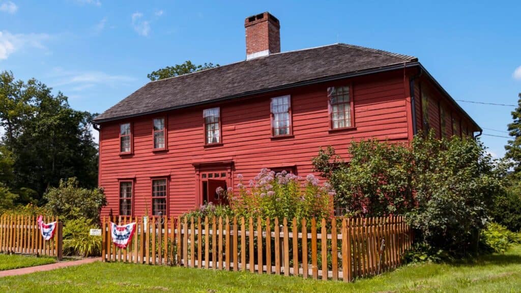 A red wooden two-story house with a brick chimney, surrounded by a brown picket fence and green garden, with patriotic banners hanging on the fence.