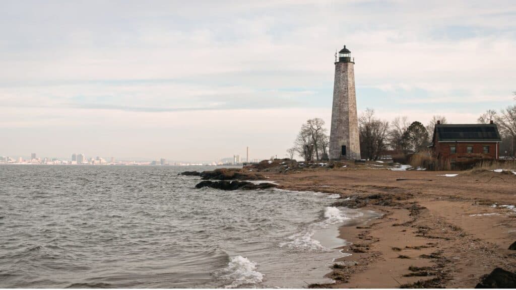 Lighthouse near a sandy shore with waves, trees, and a small building nearby; city skyline visible in the distance under a cloudy sky.