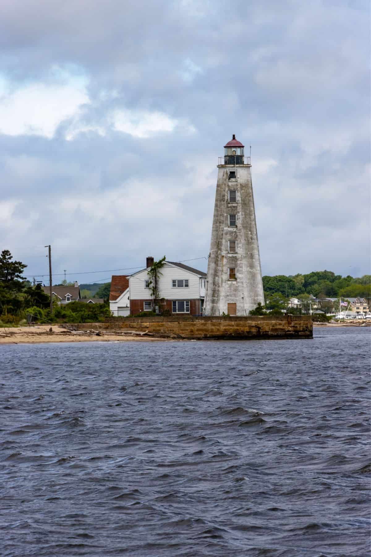 A tall, white lighthouse—one of the lighthouses you don't want to miss in Connecticut—stands beside a small house on a rocky shoreline, with trees and buildings in the background under a cloudy sky.