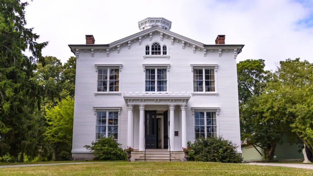 A white, two-story neoclassical house with tall windows, a columned porch, and a symmetrical facade, set against a backdrop of trees.