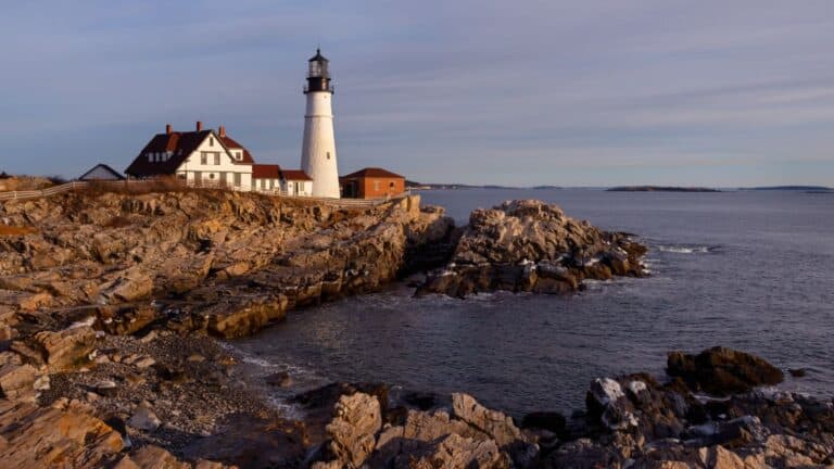 A haunted lighthouse stands on the rocky New England coastline next to a weathered house, overlooking the calm sea under a cloudy sky.