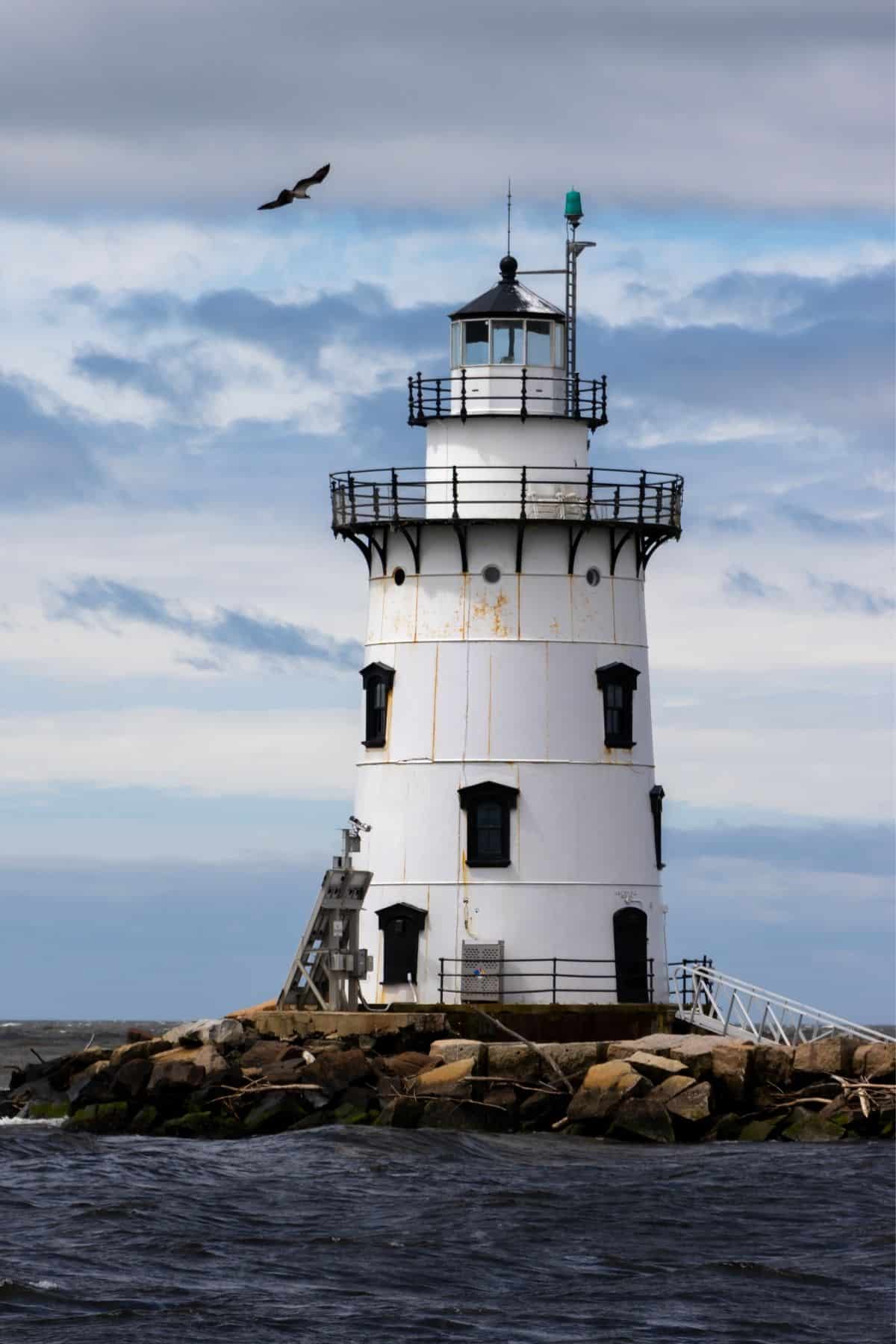 A white lighthouse with black trim stands on a rocky shore under a cloudy sky, with a bird flying above and choppy water in the foreground—one of the lighthouses you don't want to miss in Connecticut.