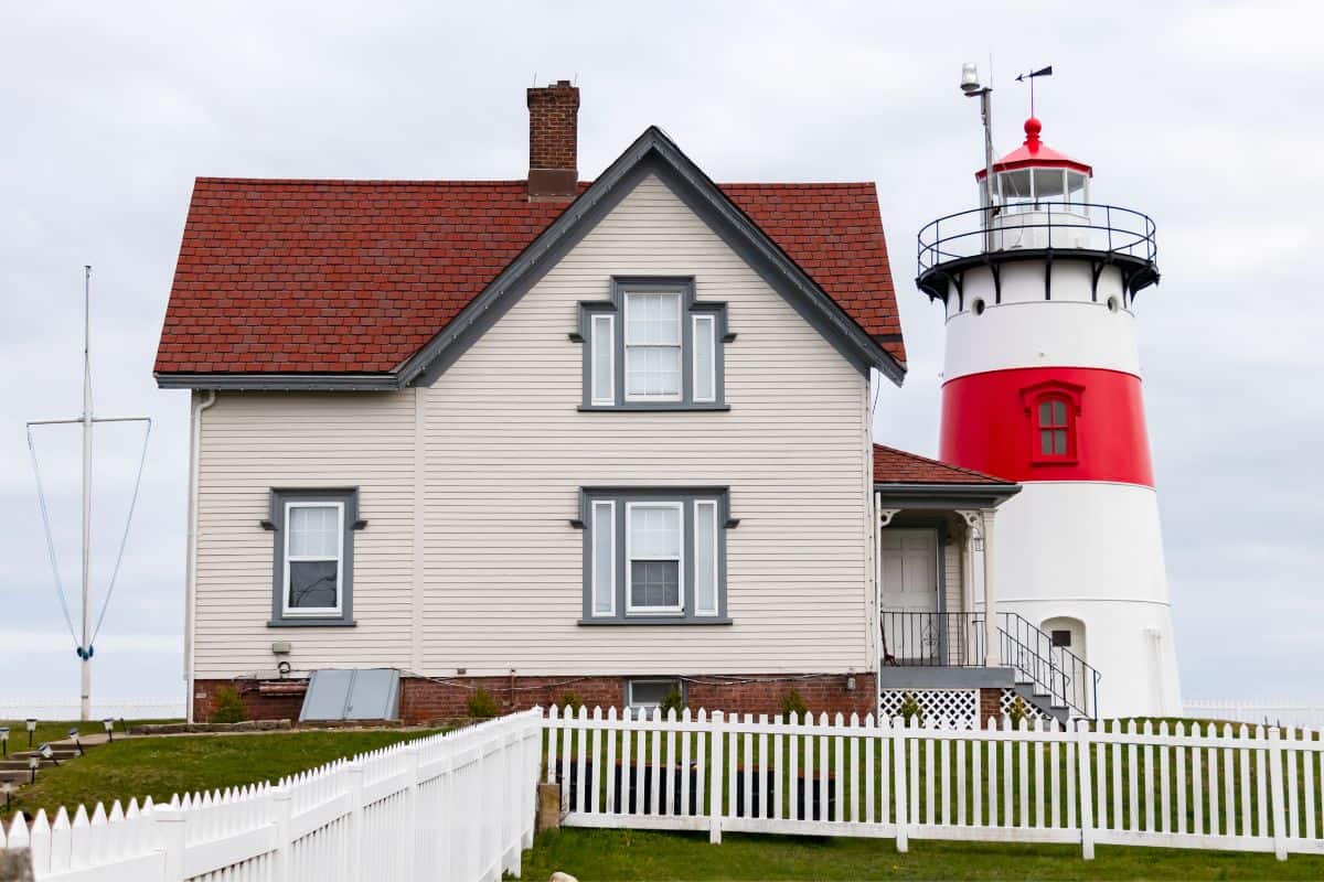 A two-story house with grey trim sits beside a red and white lighthouse, both surrounded by a white picket fence under a cloudy sky—one of those iconic lighthouses you don’t want to miss in Connecticut.