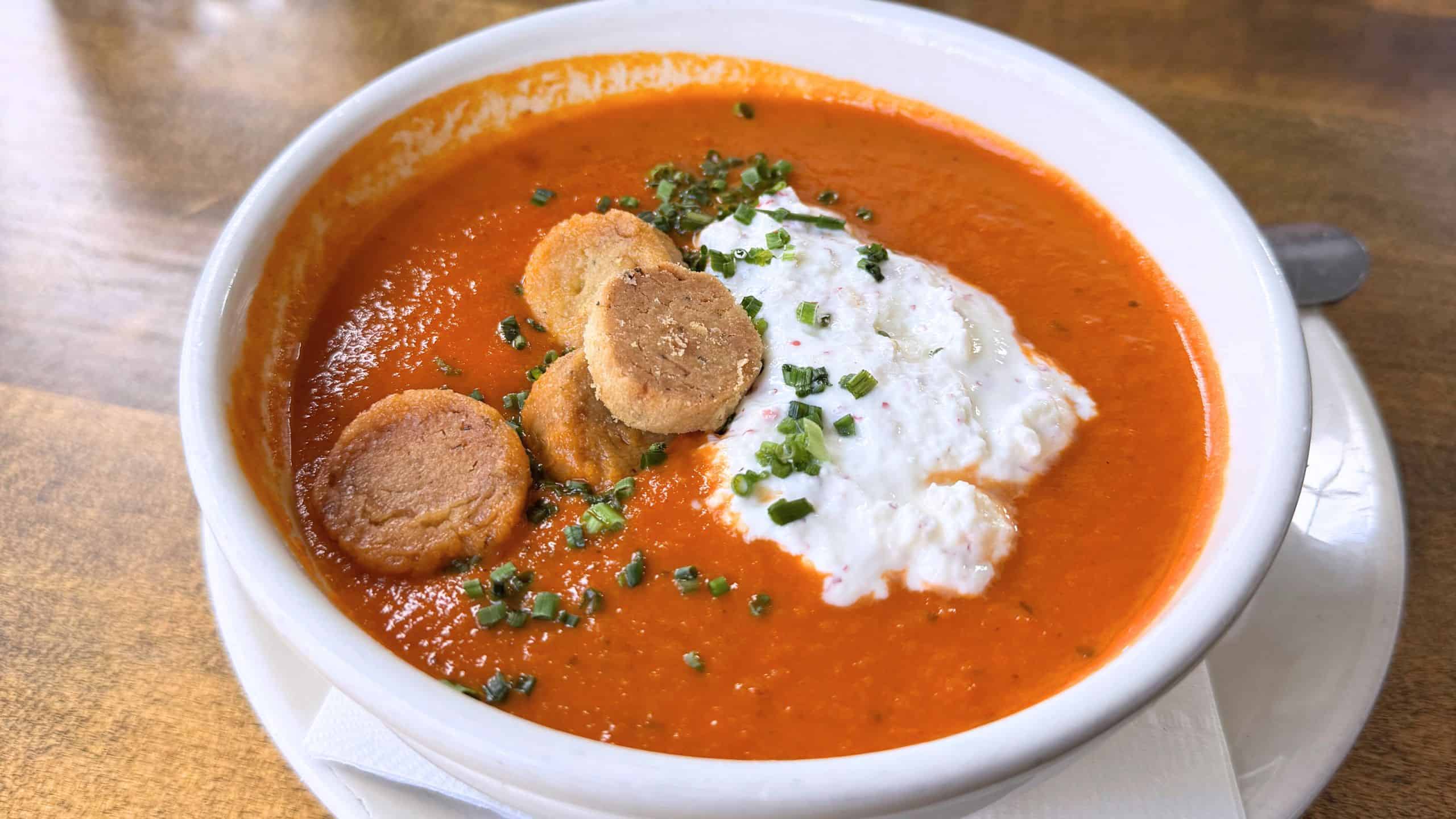 A bowl of tomato soup topped with a dollop of cream, sliced sausage, and chopped chives, served on a plate with a spoon—perfect for warming up after exploring things to do in Niantic CT.