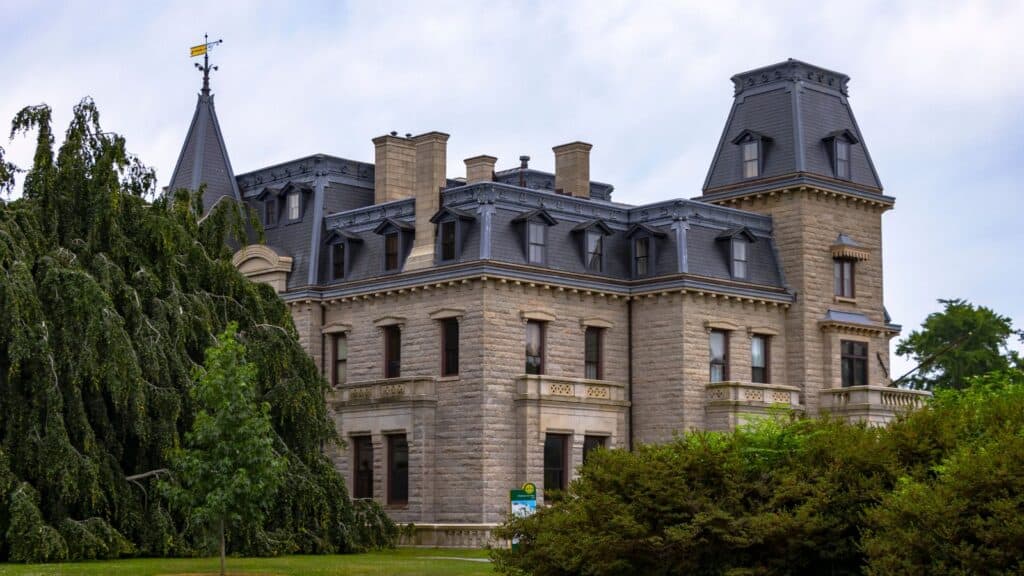 A large stone mansion with multiple windows, a slate roof, and a tower, surrounded by greenery and trees.