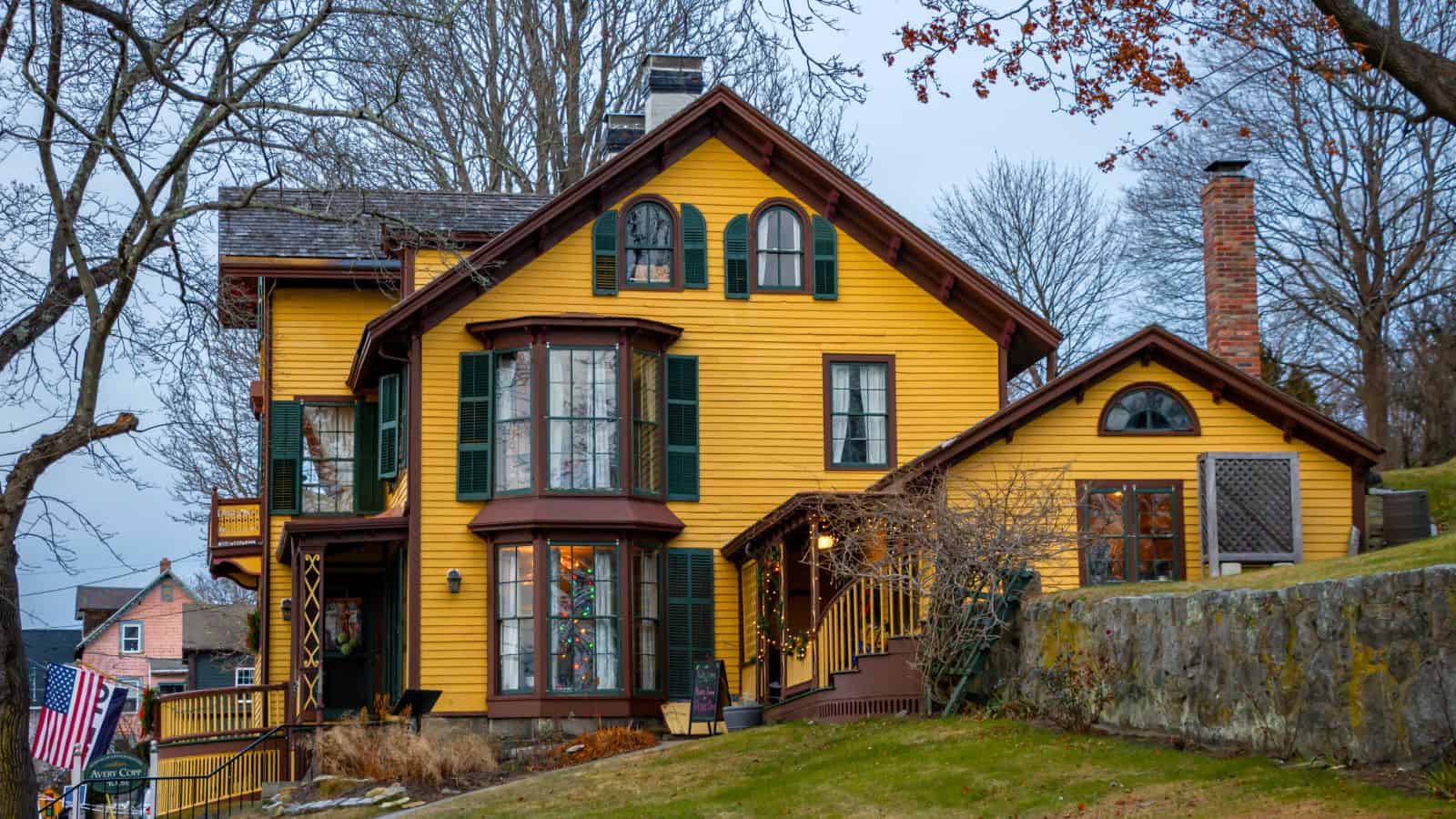 A large yellow two-story house with green shutters, a brick chimney, and a sloping lawn, surrounded by leafless trees and neighboring homes—perfect for gathering before exploring festive things to do in Connecticut this holiday season.
