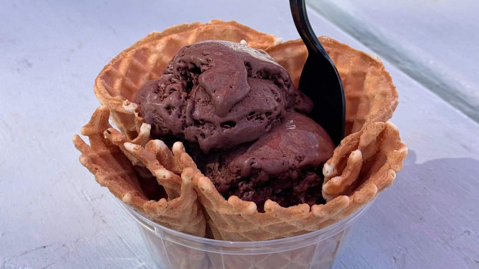 Two scoops of chocolate ice cream served in a waffle bowl with a black plastic spoon on a light-colored surface—a sweet treat to enjoy as one of the things to do in Old Saybrook.
