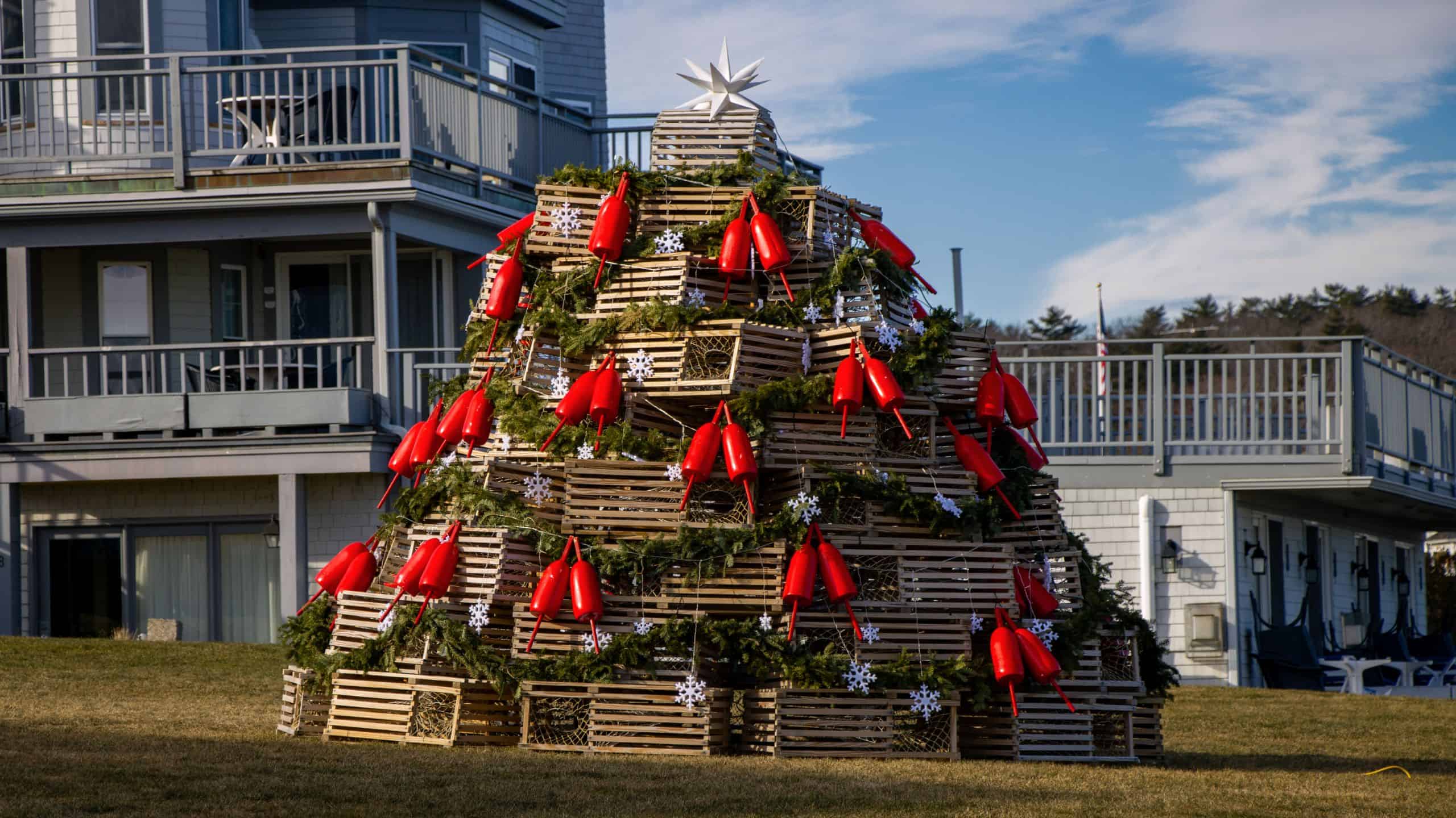 A large Christmas tree structure made of stacked wooden crates, greenery, red buoys, and a white star captures the charm of New England holidays as it stands in front of a gray building with balconies.