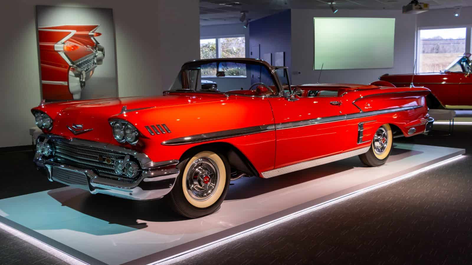 A classic red convertible car with whitewall tires is displayed on a platform in a museum setting in Newport, Rhode Island, with automotive-themed artwork in the background.