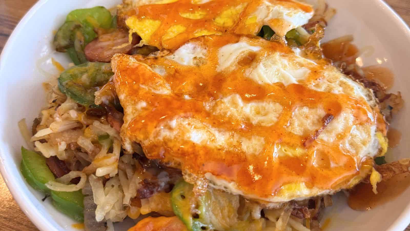 A bowl containing fried eggs topped with orange sauce, served over sautéed vegetables and shredded potatoes—a perfect start before exploring things to do in Old Saybrook.