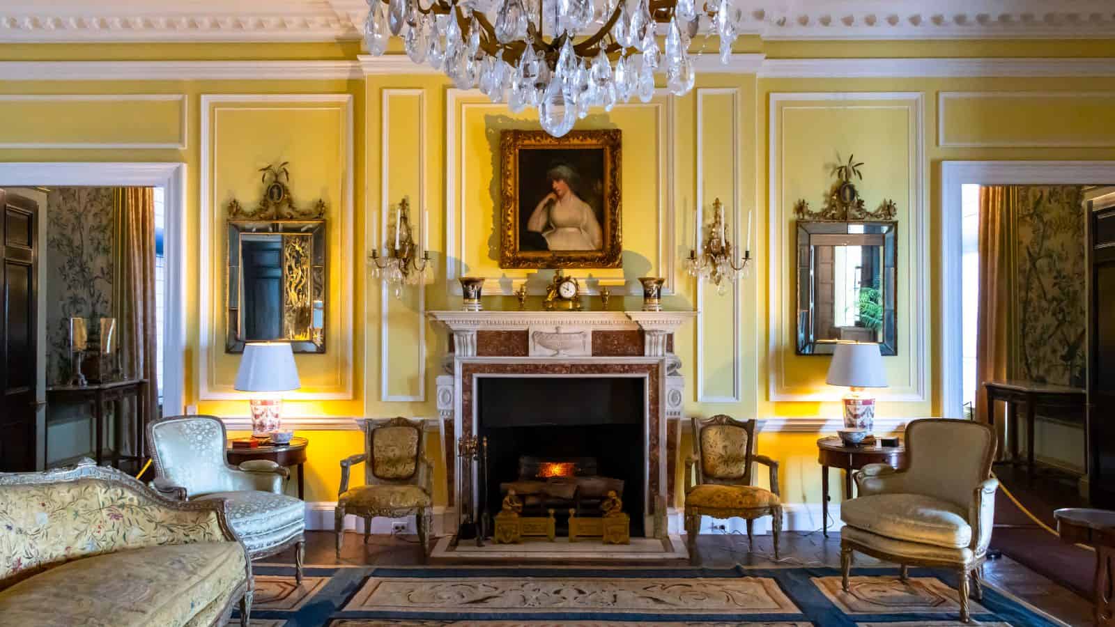 Elegant sitting room with yellow walls, a marble fireplace, a chandelier, classic furniture, two lamps, wall mirrors, and a framed portrait above the mantelpiece—reminiscent of historic Newport Rhode Island charm.