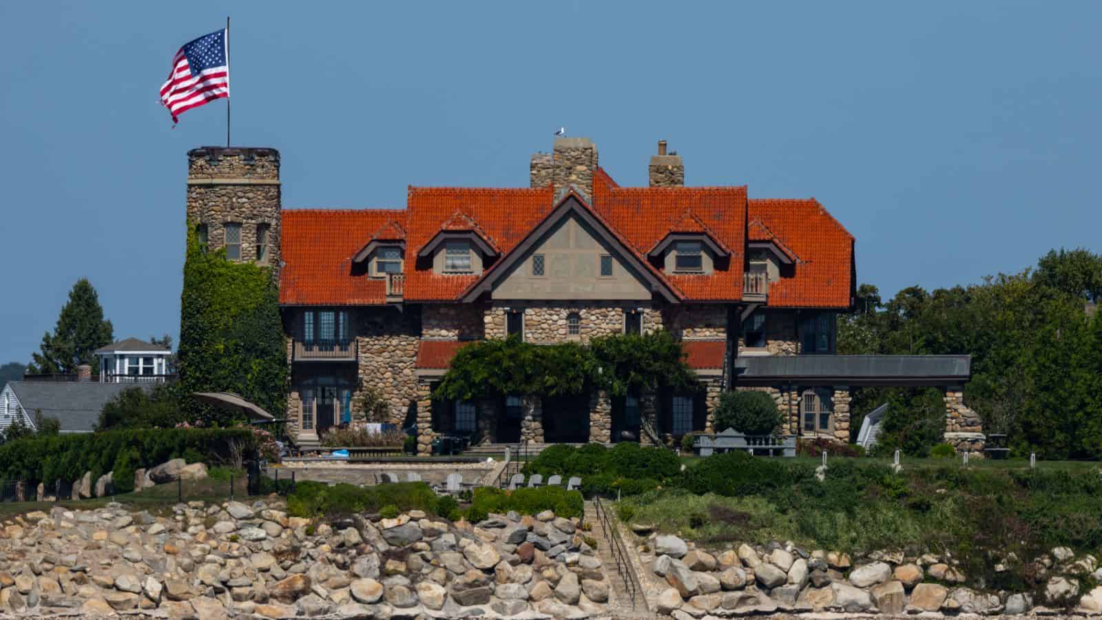 Large stone house with red roof, central tower, and American flag, situated behind a rocky shoreline and surrounded by greenery under a clear blue sky—an iconic scene that captures the charm of the area and offers inspiration for things to do in Old Saybrook.