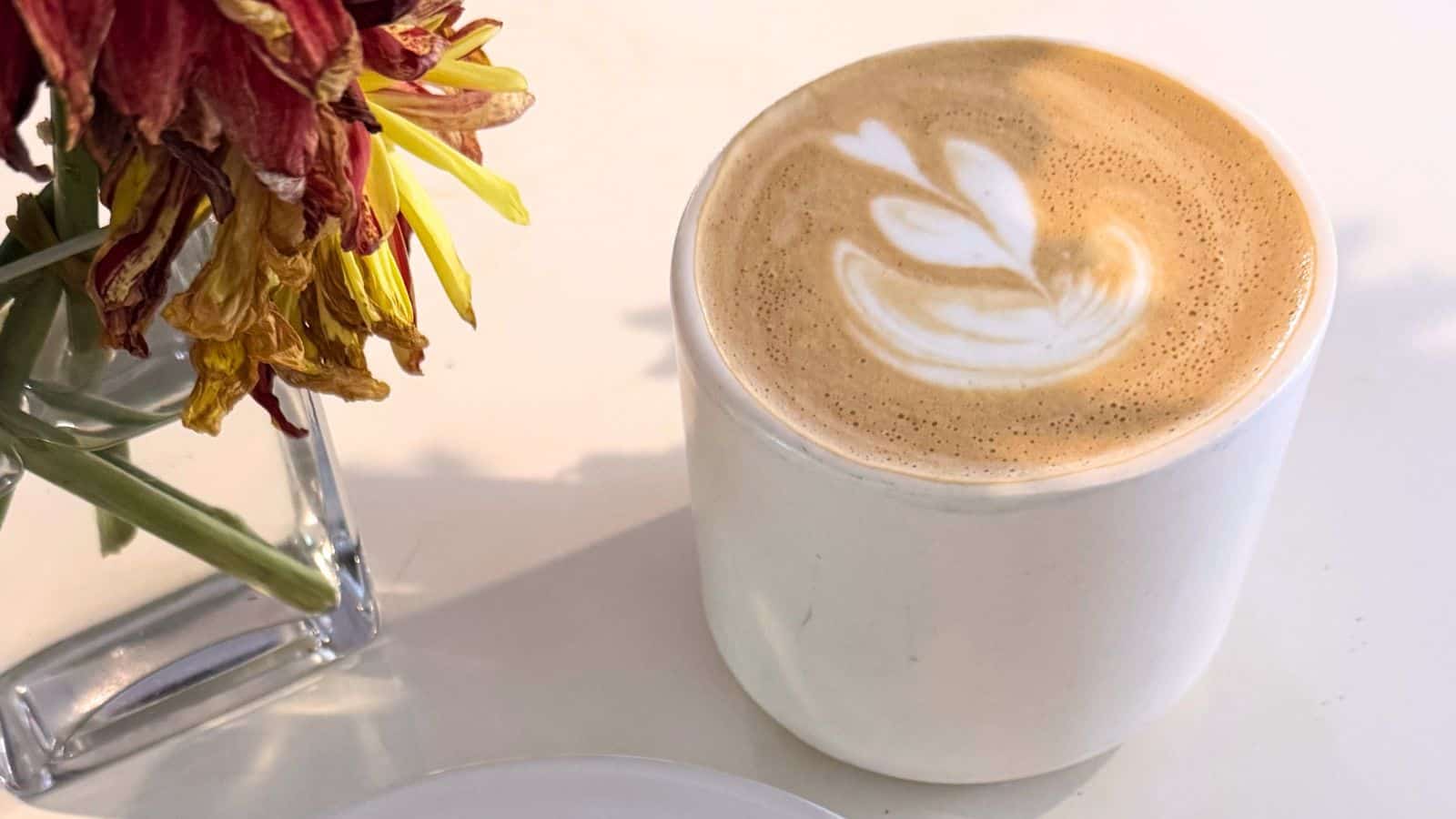 A cup of latte with a leaf-shaped latte art design sits on a white surface next to a vase of wilted flowers, evoking the cozy charm often found in Connecticut coffee shops.
