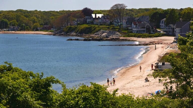 A beach with a few people walking by the shoreline and houses on a hill in the background under a sunny sky.
