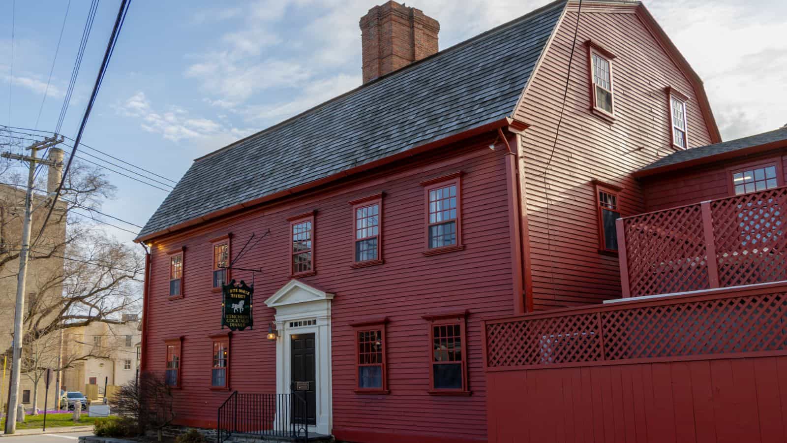 A large, red, two-story colonial-style house with a steep roof, central chimney, and white-trimmed windows, reminiscent of historic homes in Newport, Rhode Island, situated on a sunny street corner.