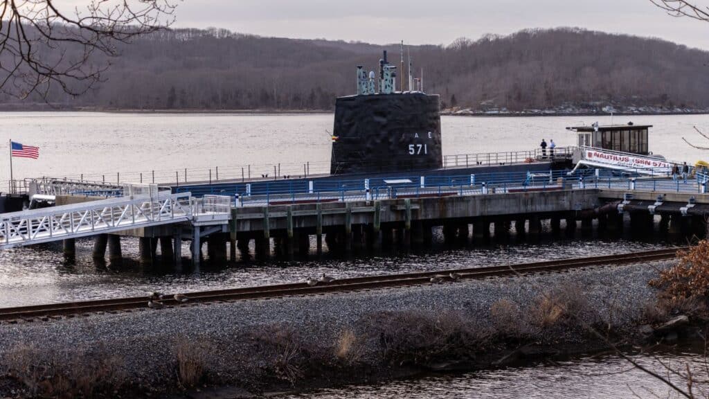 A submarine docked at a pier boasts an entrance labeled "Nautilus Submarine" in Groton, CT. Nearby, a flag gently waves, with a railway weaving alongside the wooded hills in the background.