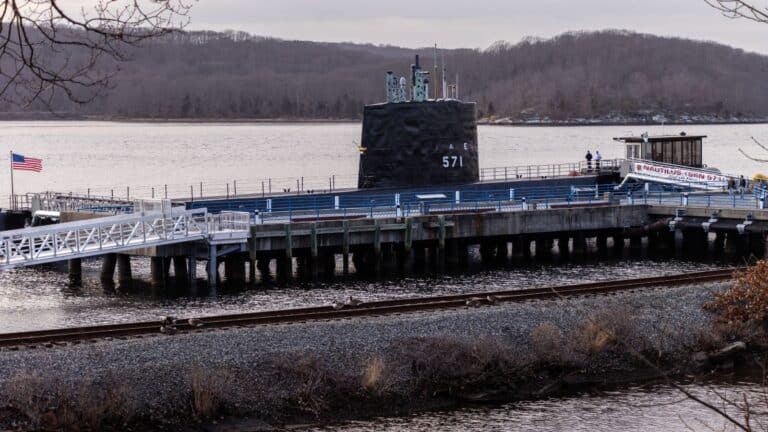 A submarine docked at a pier boasts an entrance labeled "Nautilus Submarine" in Groton, CT. Nearby, a flag gently waves, with a railway weaving alongside the wooded hills in the background.