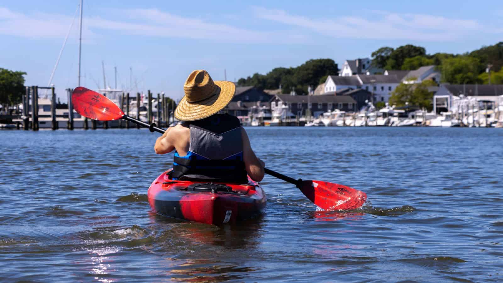Person wearing a straw hat kayaks on a calm body of water in Mystic CT, with boats and marina buildings visible in the background.