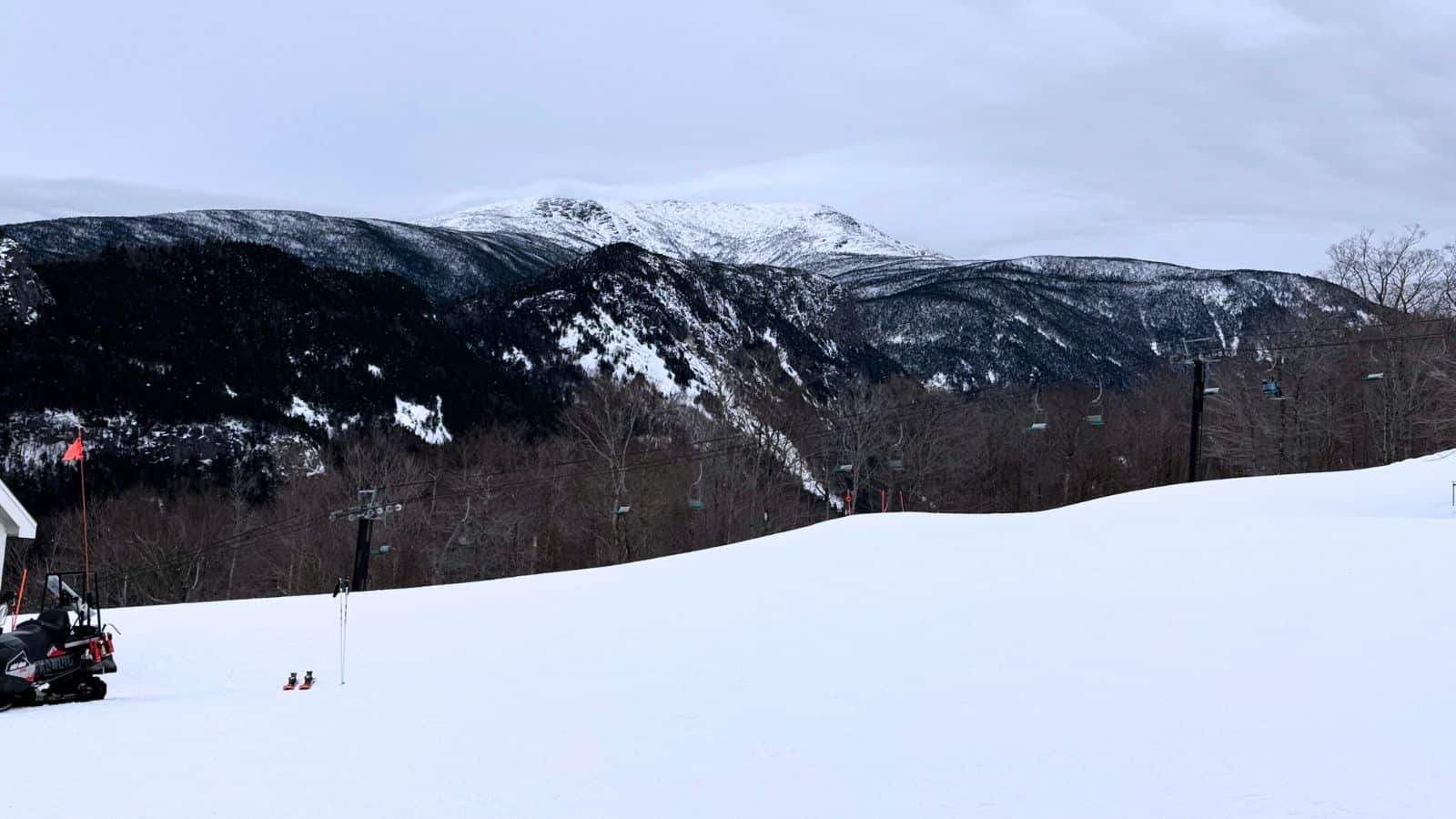 A snow-covered ski slope with ski lift chairs, bare trees, and distant mountains under a cloudy sky.