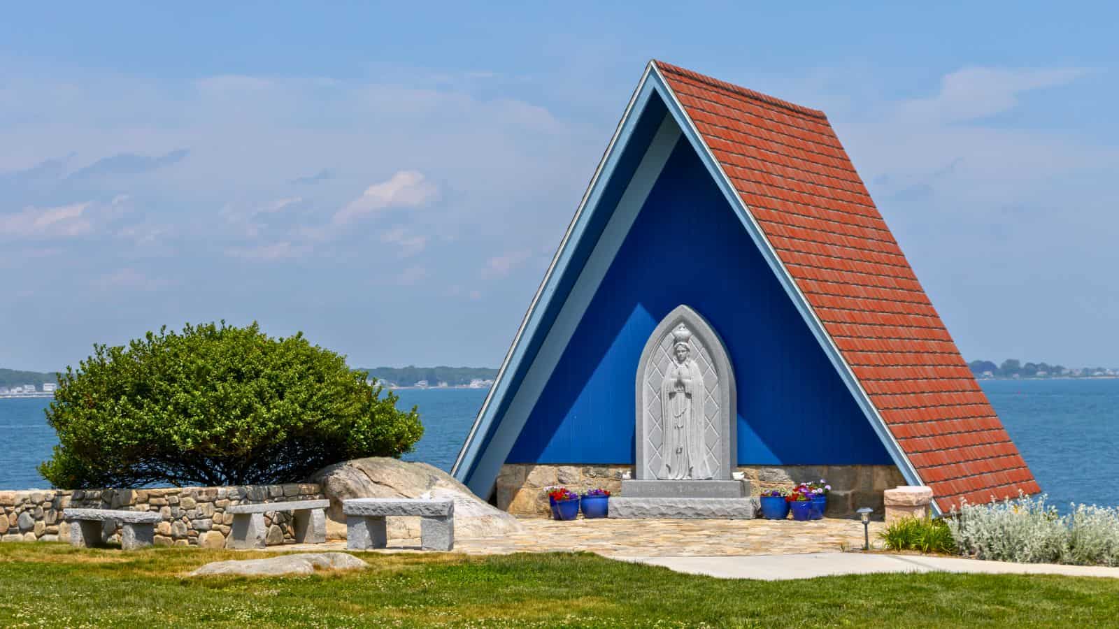 Small blue A-frame chapel with a red roof by the water in Mystic CT, featuring a statue in front, stone benches, and a stone wall surrounding the peaceful area.