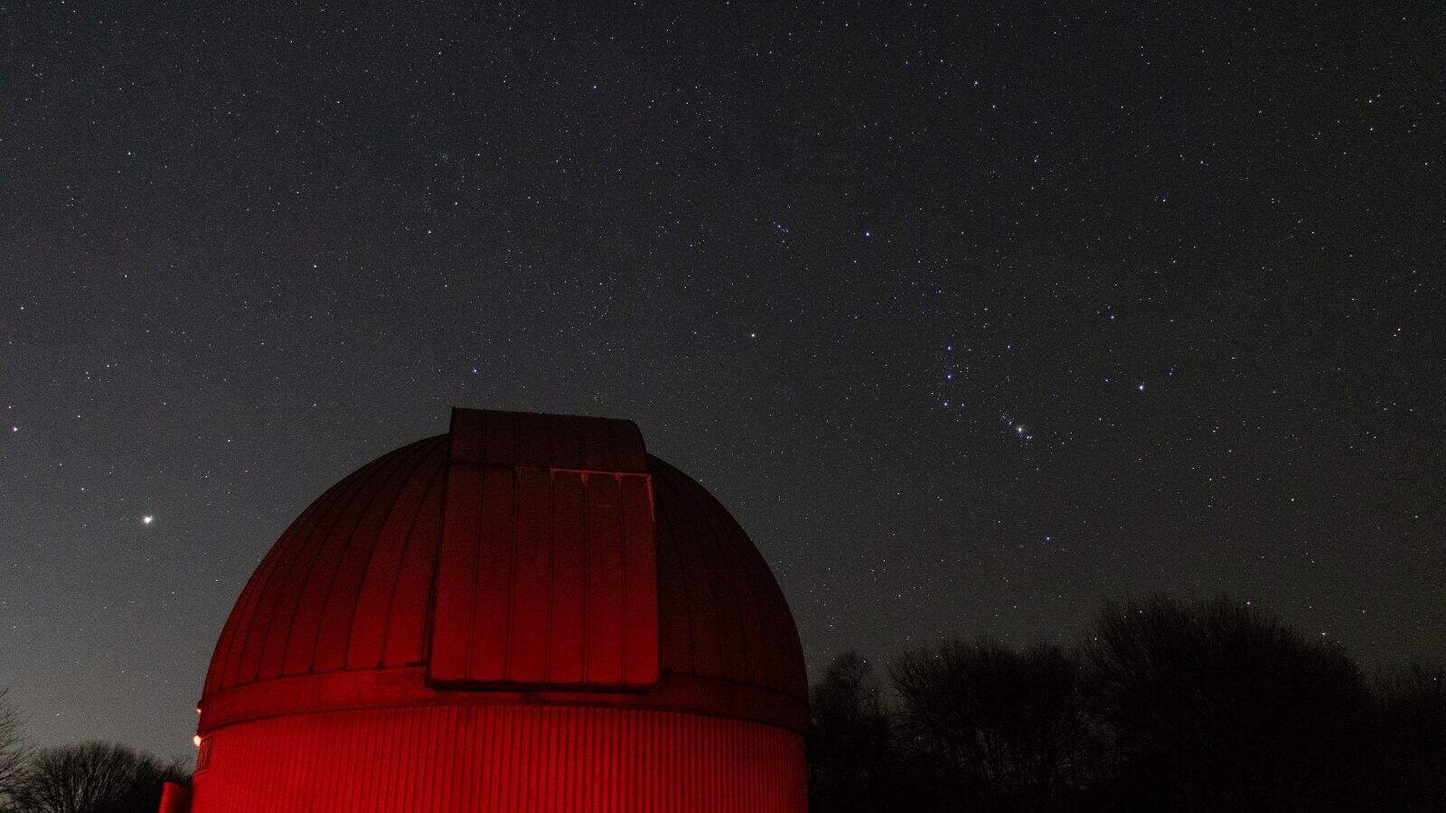 A domed observatory building in southern Rhode Island is illuminated in red light under a clear night sky filled with stars.