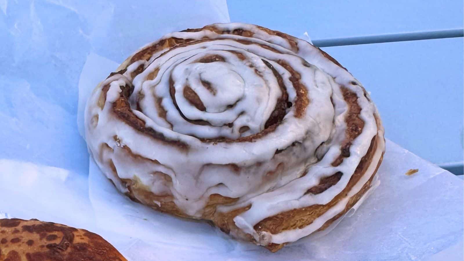 A cinnamon roll topped with white icing sits on a piece of parchment paper on a light blue table, evoking the cozy charm of Mystic CT.