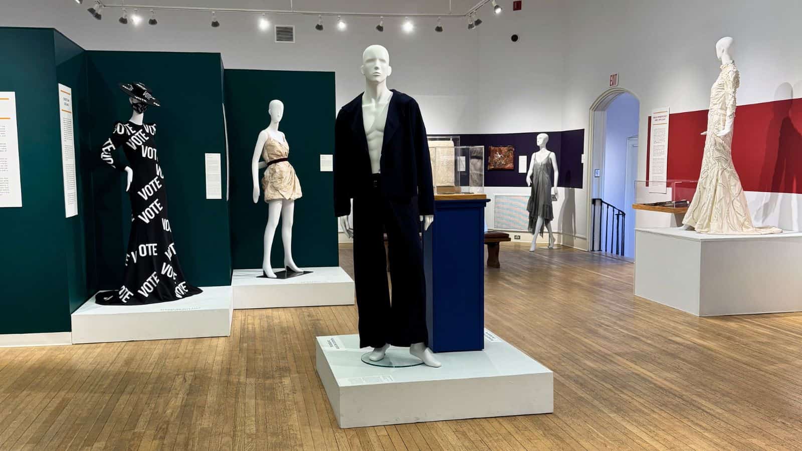 A museum exhibit in Mystic CT displays mannequins dressed in various outfits, including a black suit and a dress with "VOTE" text, on platforms in a well-lit gallery space.