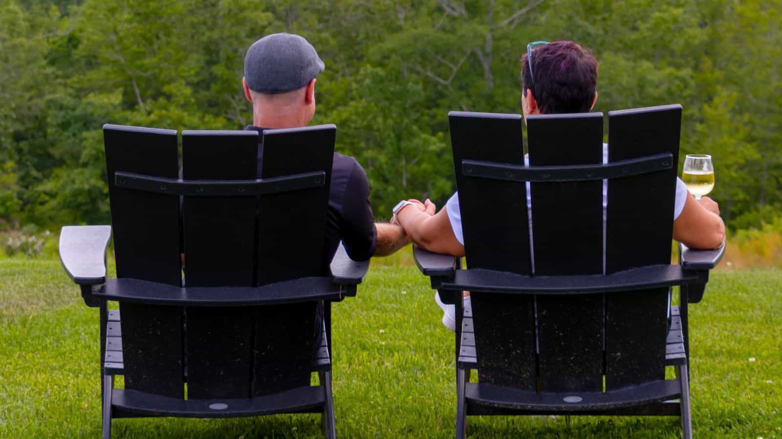 Two people sit side by side in black Adirondack chairs on grass in Mystic, CT, holding hands and relaxing outdoors; one holds a glass of white wine. Trees fill the background.