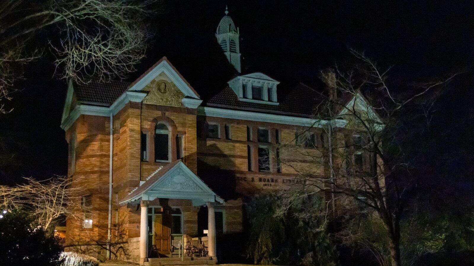 A two-story brick building in Mystic CT with a small portico, arched windows, and a cupola on the roof, photographed at night with trees partially obscuring the view.