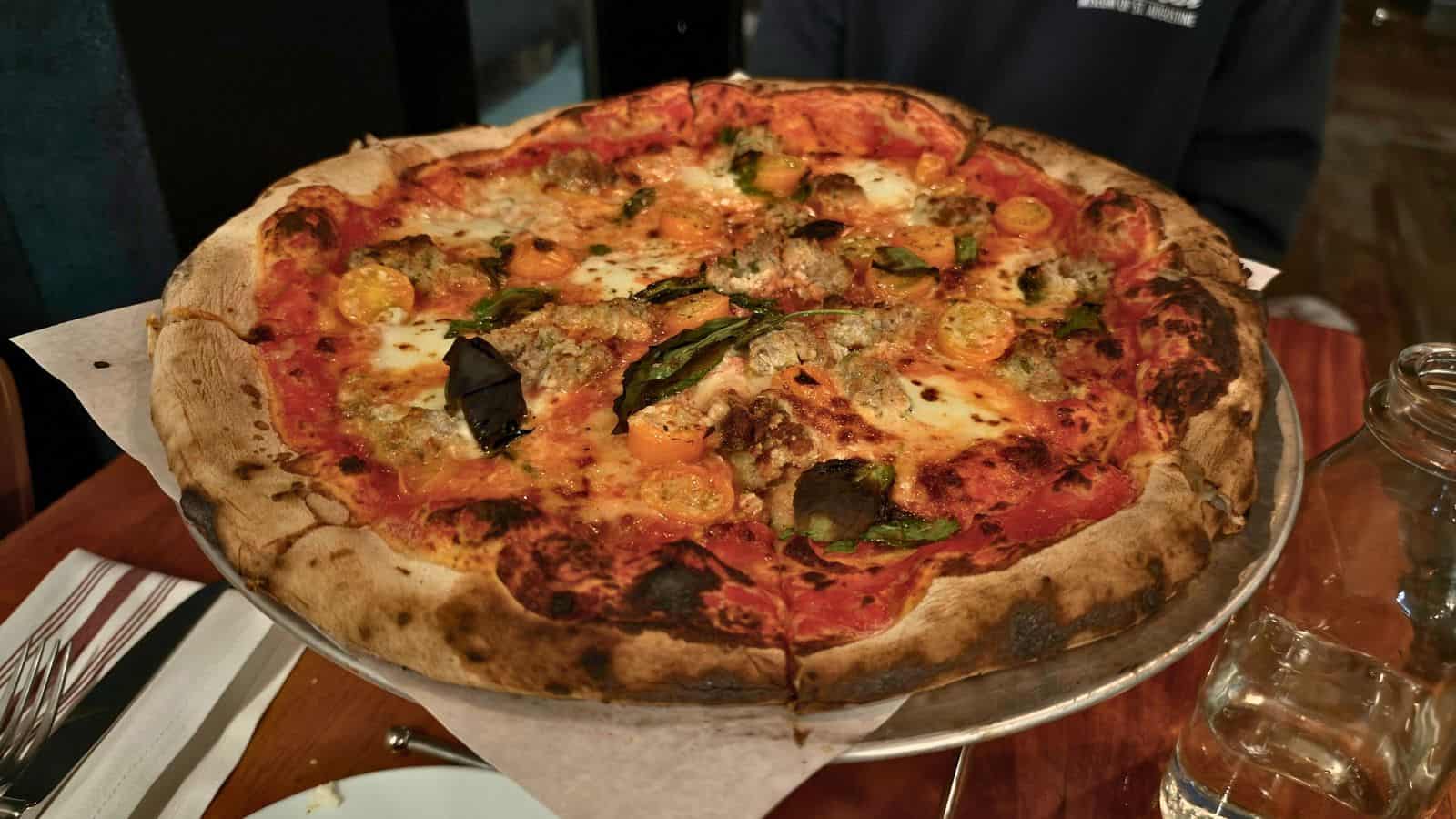 A wood-fired pizza in southeastern CT topped with tomato sauce, mozzarella, sausage, halved cherry tomatoes, and fresh basil leaves on a metal stand.