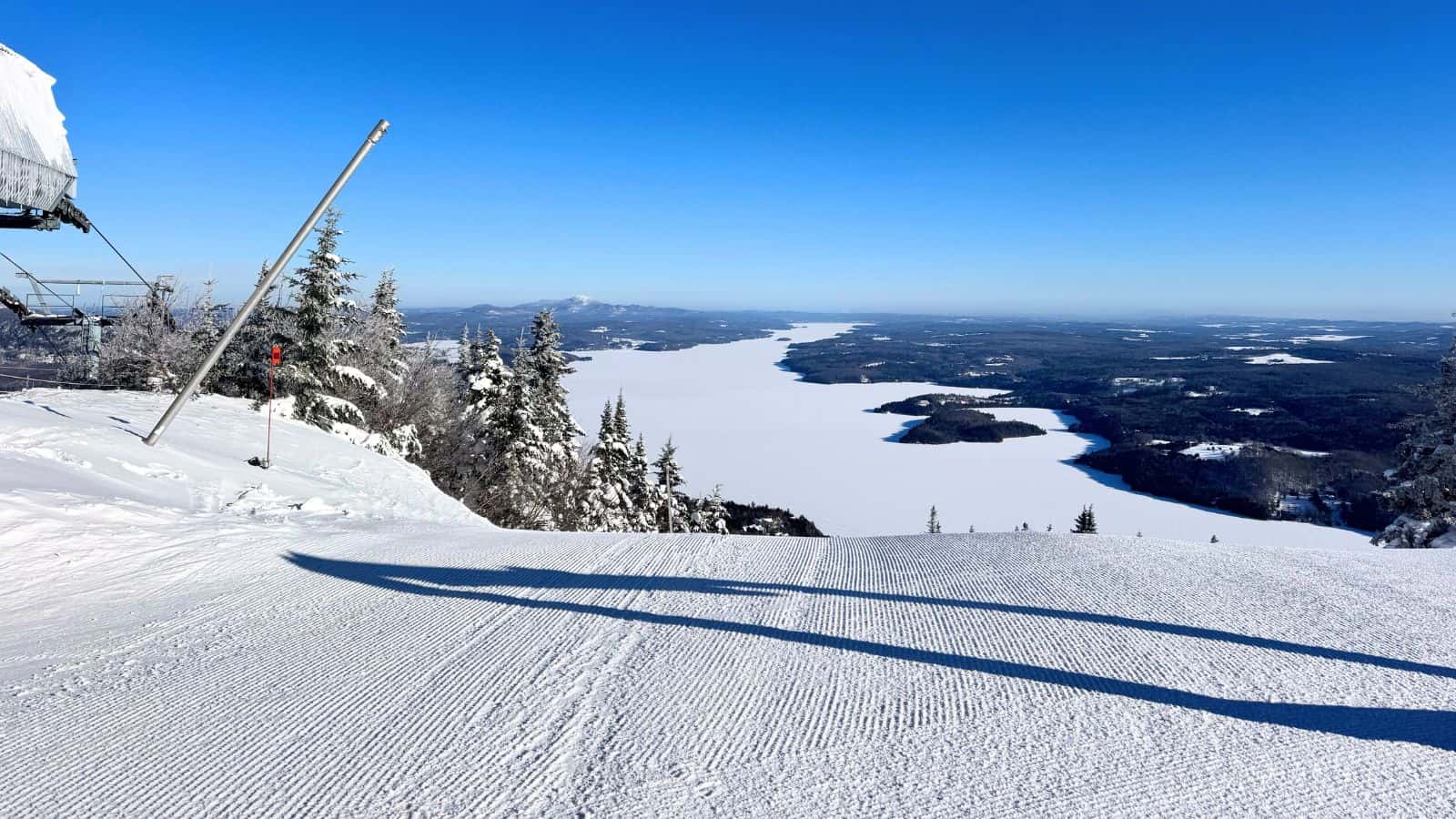 A freshly groomed ski slope overlooks a snow-covered valley and frozen lake under a clear blue sky, with trees and distant hills in the background.