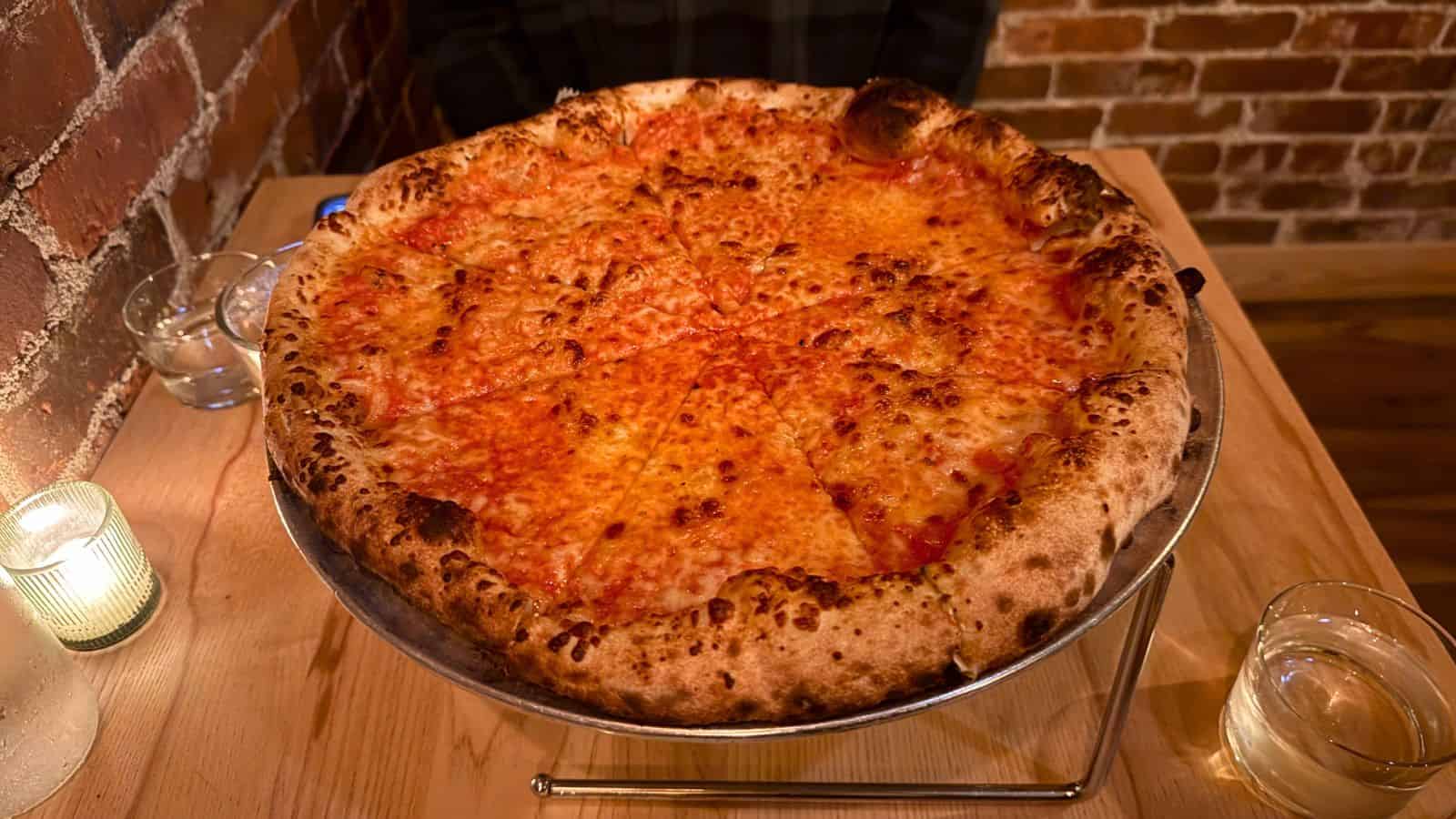 A large cheese pizza in southeastern CT with a charred crust sits on a metal stand on a wooden table, next to a glass of water and a candle, against a brick wall background.