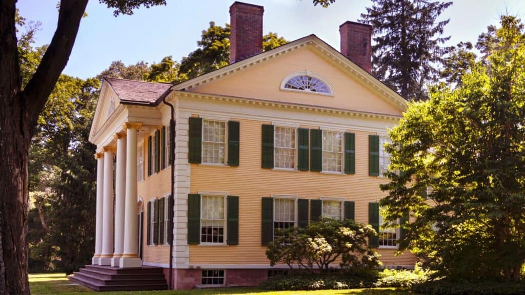 Yellow two-story house with green shutters, white columns, and a gabled roof, surrounded by trees and greenery.