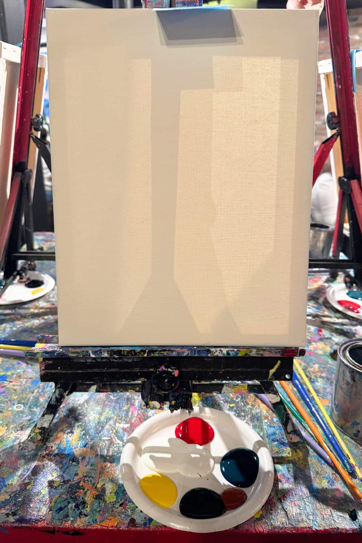 An easel holds a blank canvas. Below is a palette with dabs of red, yellow, blue, and black paint. The setup is on a colorful, splattered table.